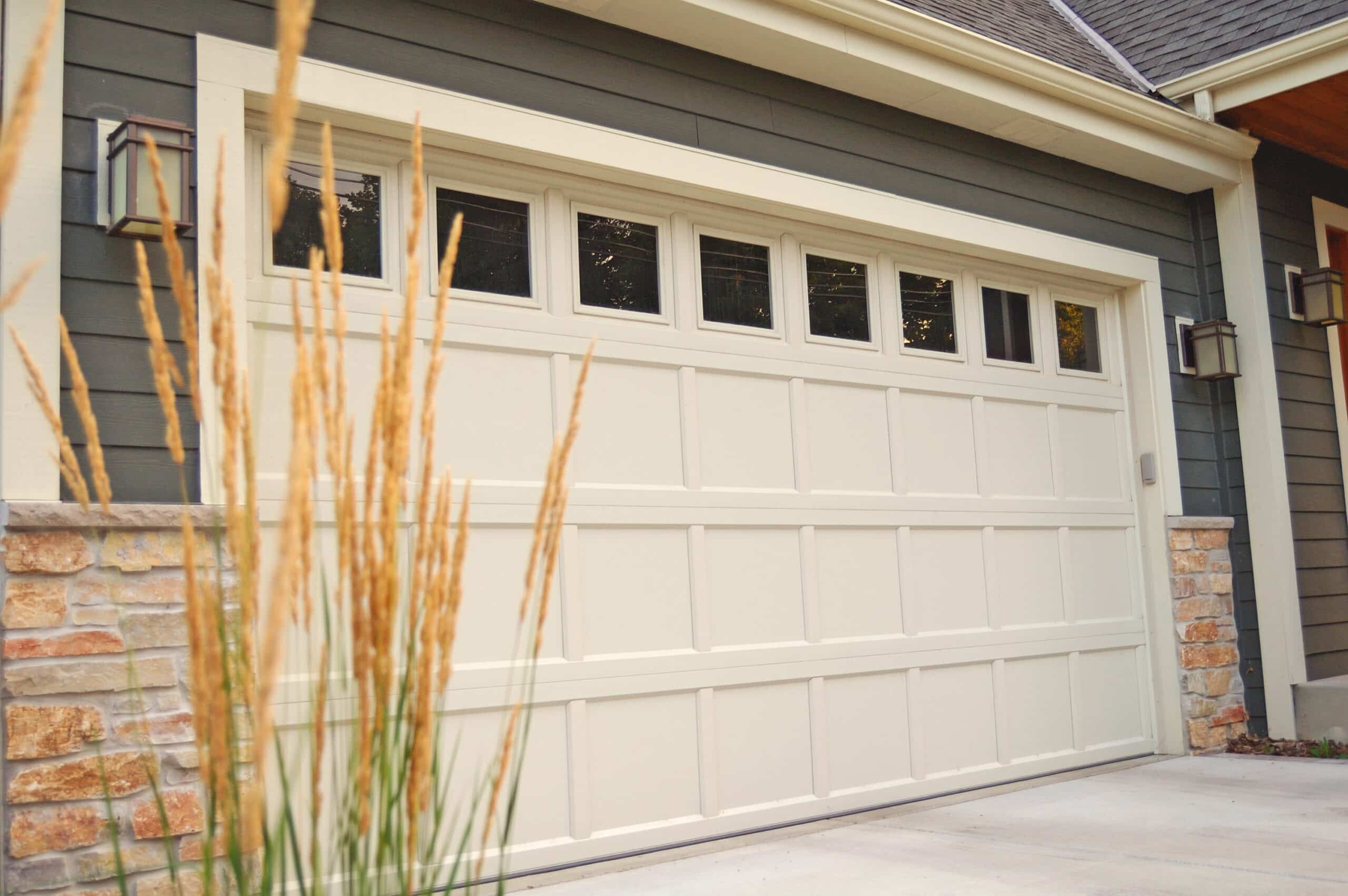 Gray sectional garage door with a row of small rectangular windows above, stone veneer on the sides, and dusk-style exterior lighting. (informative)