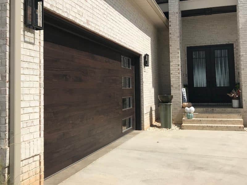 Wide view of a brick house entry: a large dark wood garage door on the left and a black front door with glass panels on the right, with steps and potted plants by the doorway.