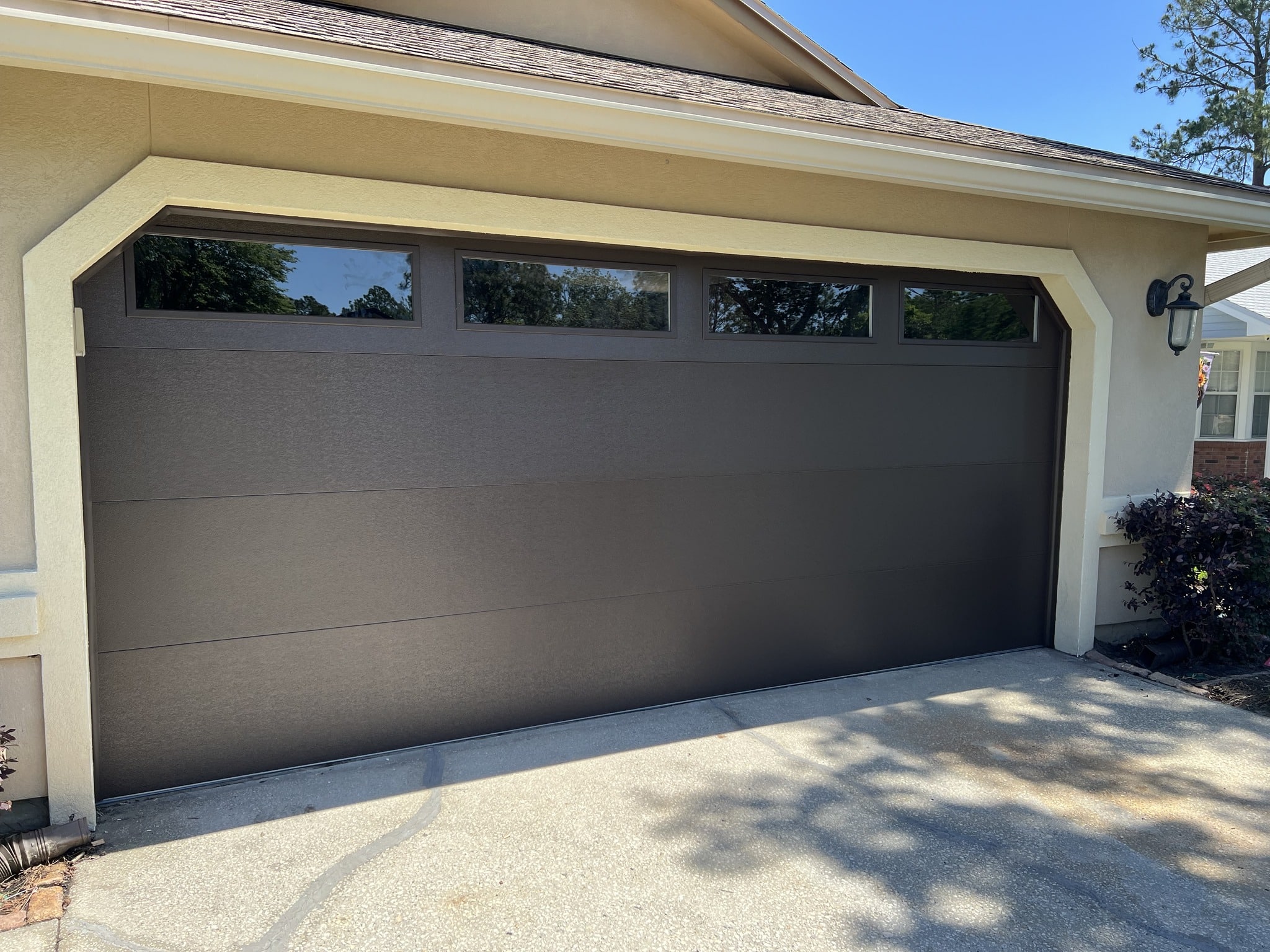 Dark gray sectional garage door with four horizontal rectangular windows, beige stucco home, and a lantern-style exterior light on the right under a blue sky.
