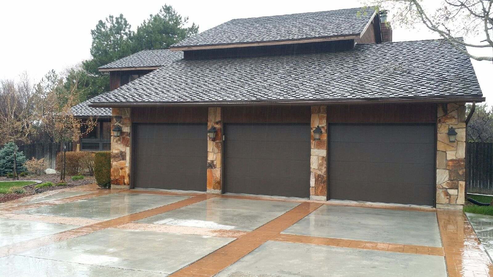 Three-car garage with dark brown doors, stone columns, and a slate roof on a wet concrete driveway.