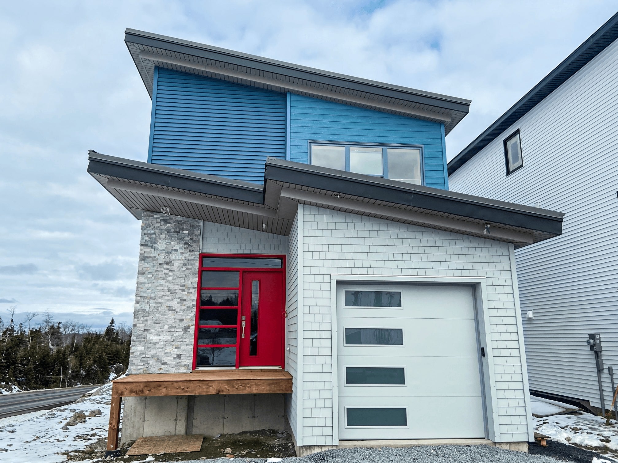 Modern two-story house with blue siding, red front door, and a white garage door; stone pillar at entrance and wooden steps in snowy landscape.