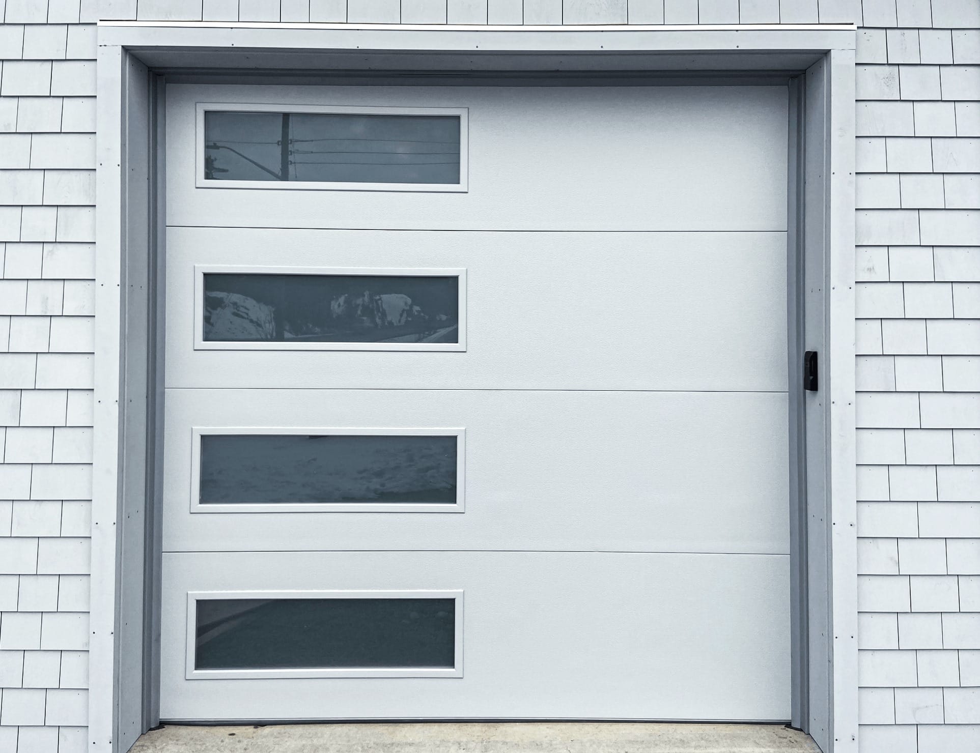 Light gray sectional garage door with three horizontal rectangular windows, surrounded by white clapboard siding and metal trim.