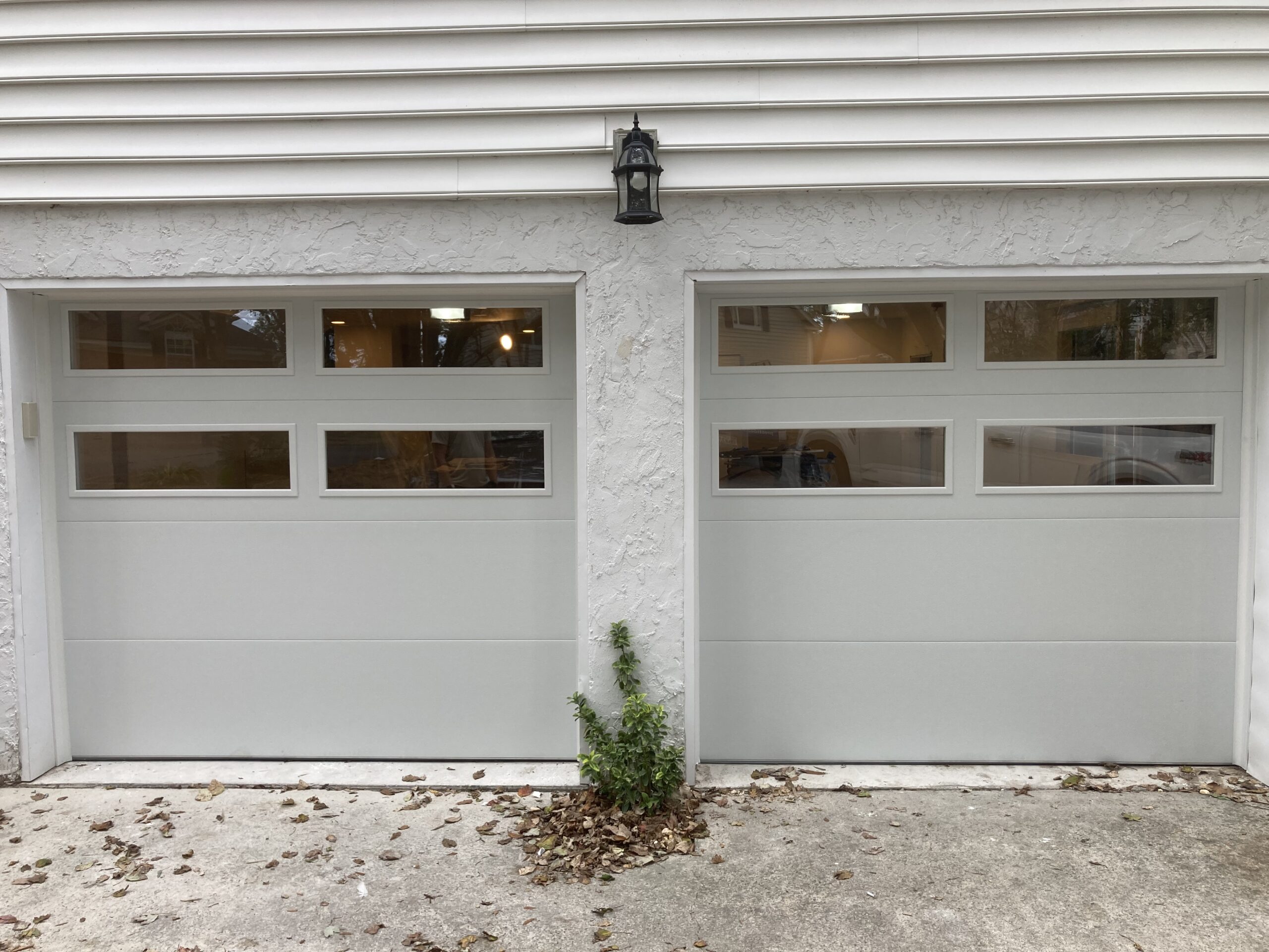 Two light-gray garage doors separated by a stucco column, with a black lantern centered above and fallen leaves on the driveway.