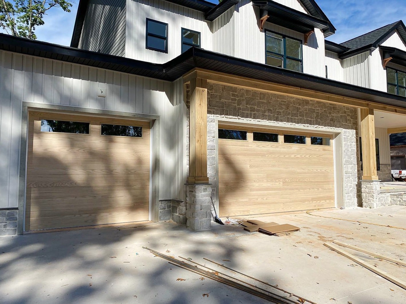 New modern house with three beige wood-paneled garage doors, stone pillars, and light siding under construction in bright daylight