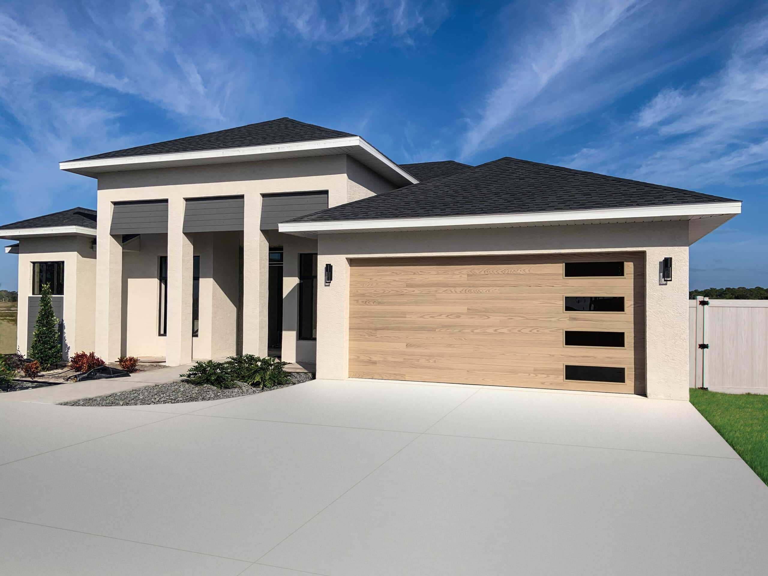 Front view of a modern beige suburban house with a wooden garage door and dark roof under a blue sky.